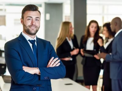 Caucasian businessman leader looking at camera in modern office with multi-ethnic businesspeople working at the background. Teamwork concept. Young man with beard wearing blue suit.