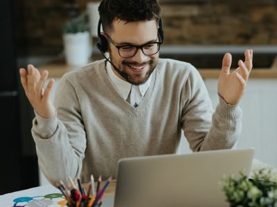 Happy businessman with headphones talking to someone while making video call over laptop from home.